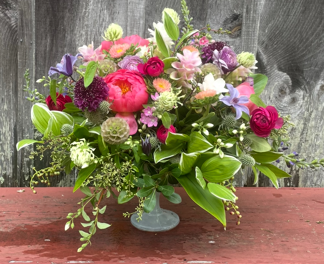 Low compote of coral peony, ranunculus, scabiosa, clematis and trailing greenery against a weathered fence