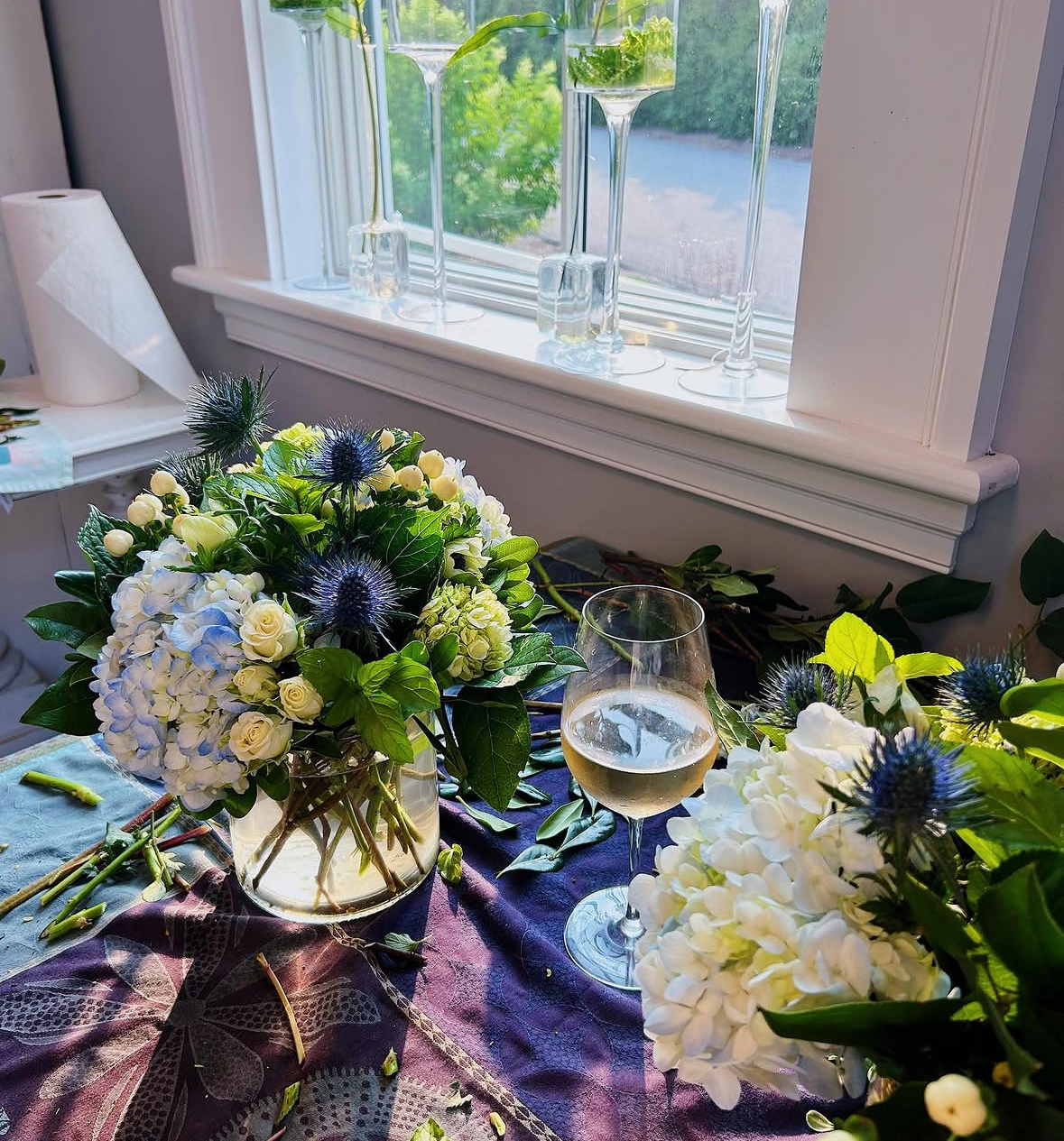 White hydrangea and blue thistle arrangement in progress beside a glass of wine